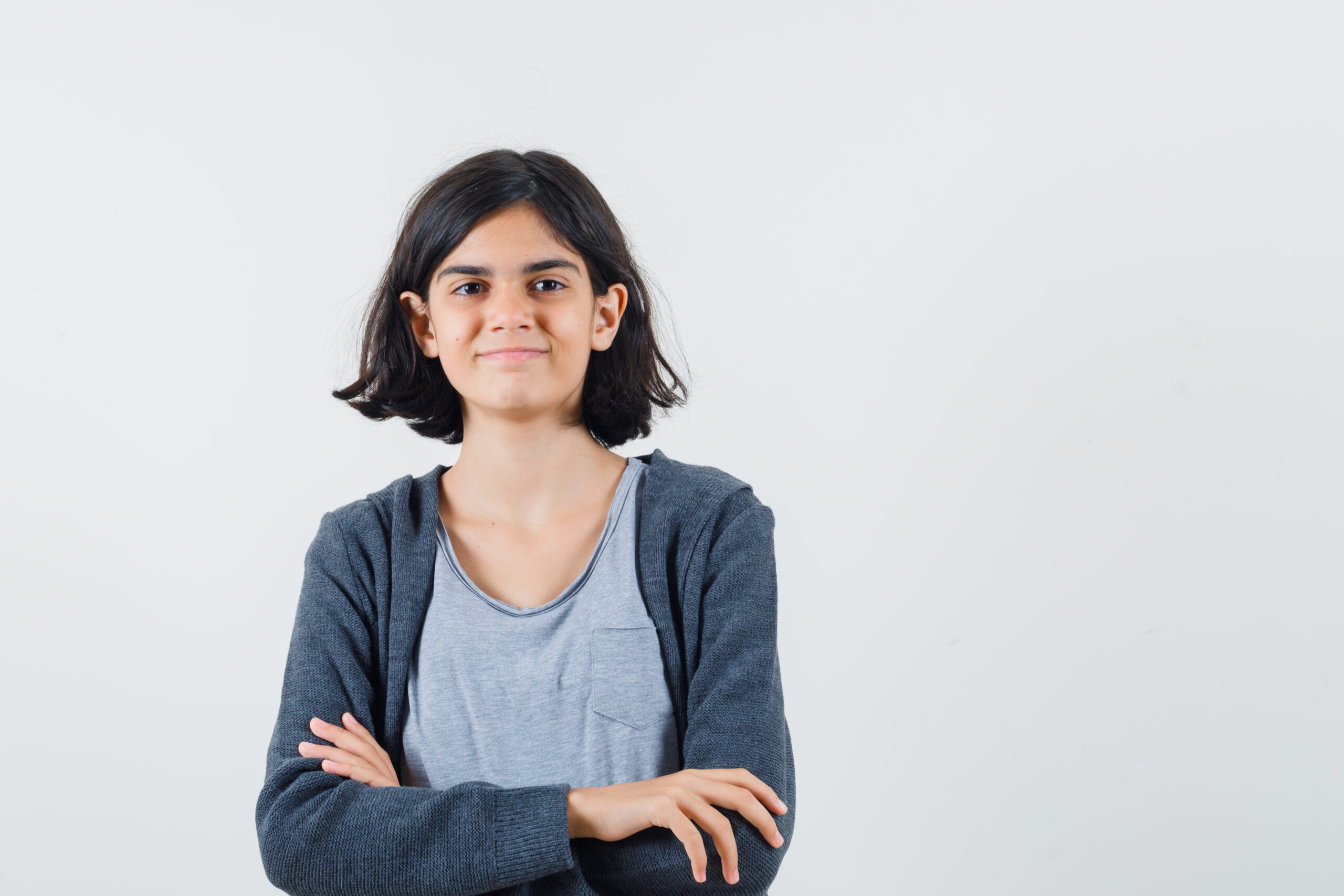 young girl standing arms crossed in light gray t shirt and dark grey zip front hoodie and looking cute , front view.